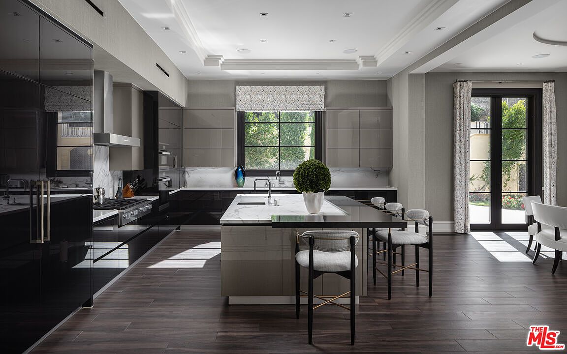 This is a modern kitchen with sleek, glossy cabinetry in dark and light gray tones, complemented by marble countertops and backsplash. A large island with a waterfall edge provides ample workspace and seating, while a window above the sink offers natural light and a view of the outdoors. The dark wood flooring adds warmth and sophistication to the space.