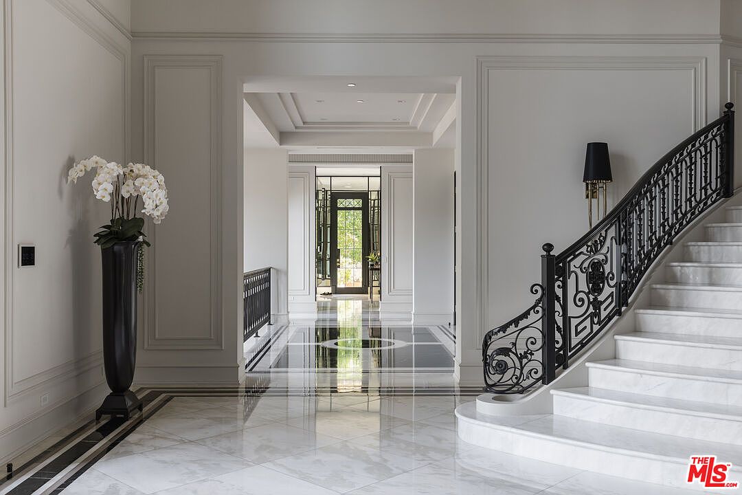 This interior shot showcases a grand hallway with marble flooring and a striking black and white design. A staircase with ornate black iron railings adds an elegant touch, while a long hallway leads to a door at the end, creating a sense of depth and spaciousness. The walls are white with decorative molding, and a tall black vase with white flowers sits to the left.