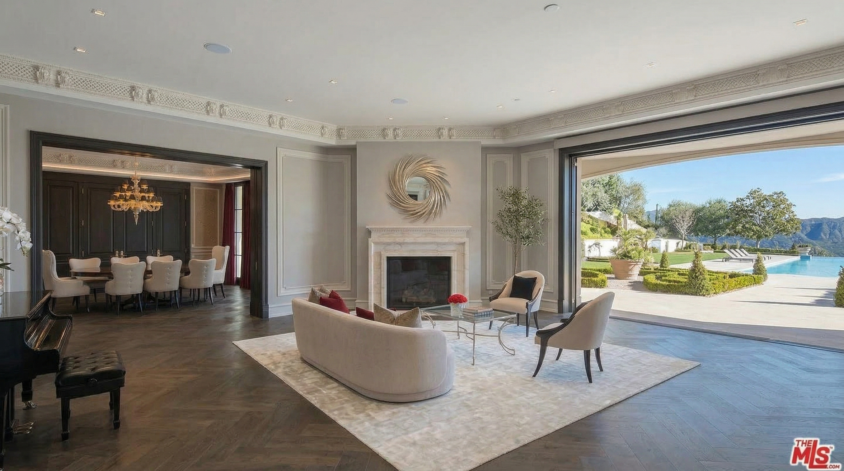 This elegant interior living space features sophisticated herringbone hardwood flooring and a central fireplace adorned with a modern sunburst mirror. A wide, seamless opening connects the room to an expansive outdoor patio and pool area, emphasizing a luxurious indoor-outdoor lifestyle. The room is further complemented by a graceful curved sofa and an adjacent, refined dining area.