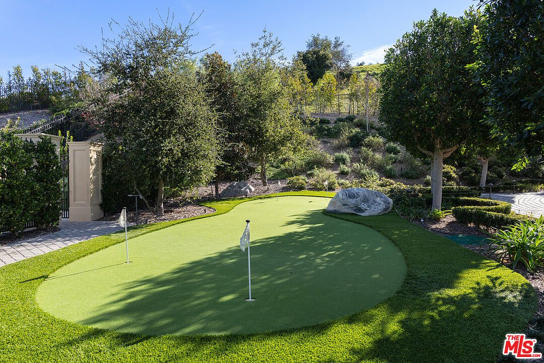This image showcases a meticulously maintained backyard putting green, complete with flags and surrounded by lush landscaping. Mature trees and manicured shrubs frame the green, creating a serene and private atmosphere. A large rock adds a natural element to the scene, enhancing the overall aesthetic appeal.