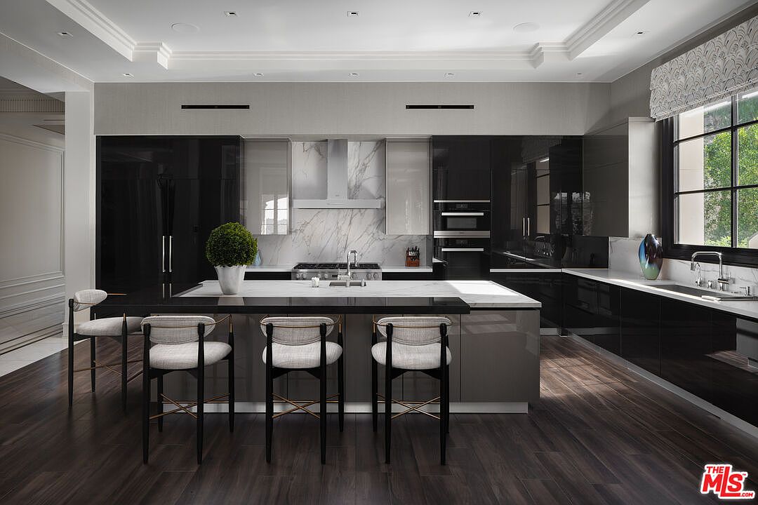 This is a modern kitchen with sleek, dark cabinetry and a contrasting white marble backsplash and countertops. A large island with bar seating anchors the space, while stainless steel appliances and a minimalist range hood add to the contemporary aesthetic. The dark wood flooring provides a warm contrast to the cool tones of the cabinetry and countertops.