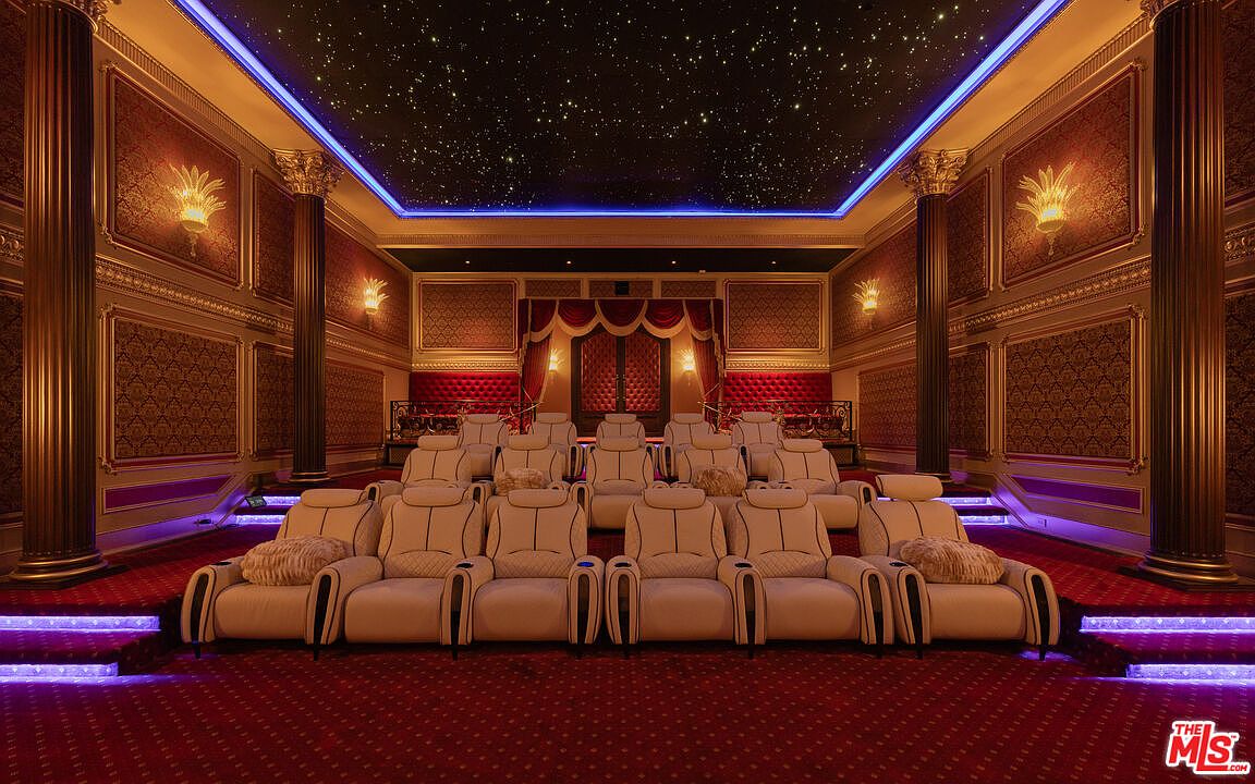 This is a luxurious home theater featuring tiered seating with plush, cream-colored reclining chairs, each equipped with cup holders. The room is adorned with ornate, gold-accented walls and columns, a red carpet, and a ceiling designed to mimic a starry night sky with blue accent lighting. The overall impression is one of opulence and comfort, perfect for an immersive cinematic experience.