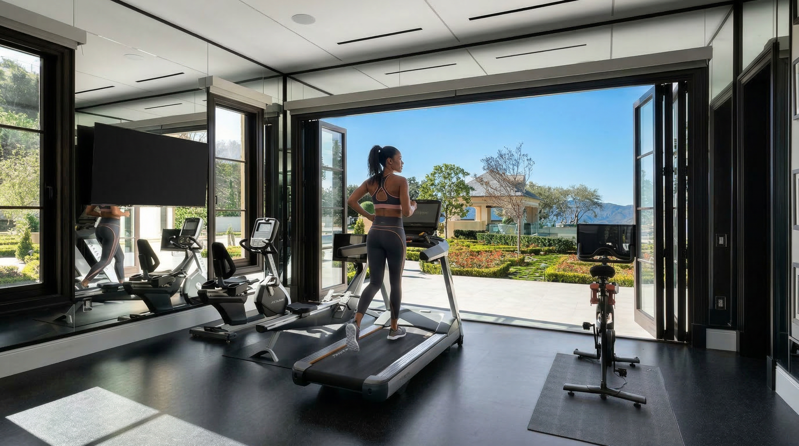 This luxurious, high-end home gym features cutting-edge exercise equipment, including a treadmill and stationary bike, set against a backdrop of floor-to-ceiling accordion glass doors. The open glass wall provides a seamless transition to a manicured garden landscape, flooding the room with natural light. The sophisticated design includes dark flooring and built-in mirror reflections, creating an expansive, professional workout environment.