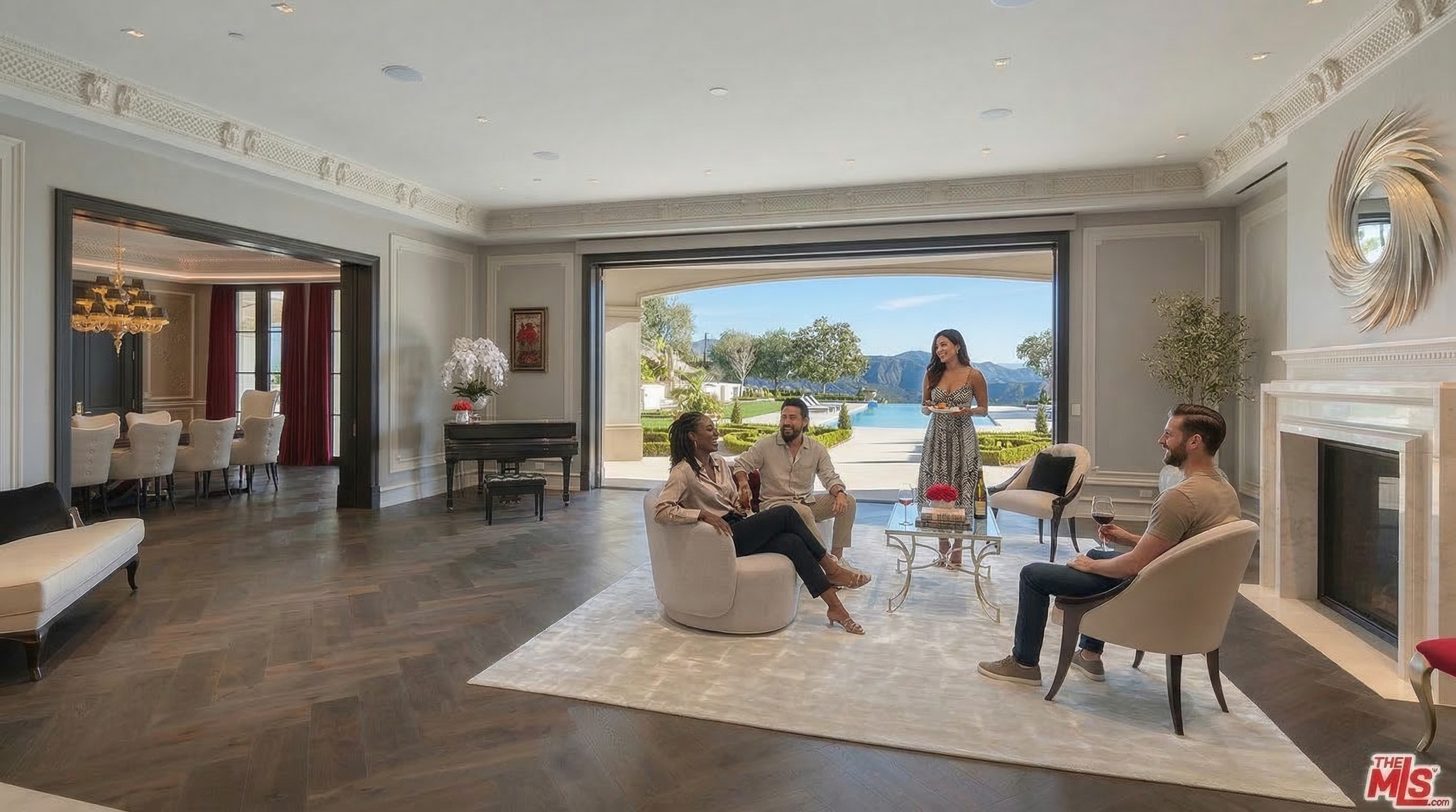 This elegant and spacious living room features luxurious hardwood floors in a herringbone pattern, intricate crown molding, and a grand fireplace. Large, open pocket doors seamlessly connect the interior space to an outdoor patio with stunning mountain and pool views, creating an ideal layout for entertaining. The room is styled with contemporary neutral-toned furniture, creating a sophisticated and inviting atmosphere.