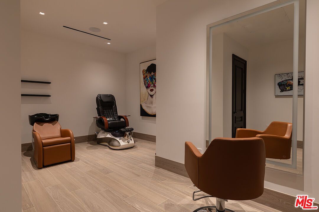 This interior shot showcases a salon or spa room, featuring a shampoo station with a brown leather chair, a black massage chair on a white base, and a brown leather salon chair reflected in a large mirror. The room has light wood flooring and neutral-colored walls, creating a clean and relaxing atmosphere, enhanced by modern lighting and artwork.