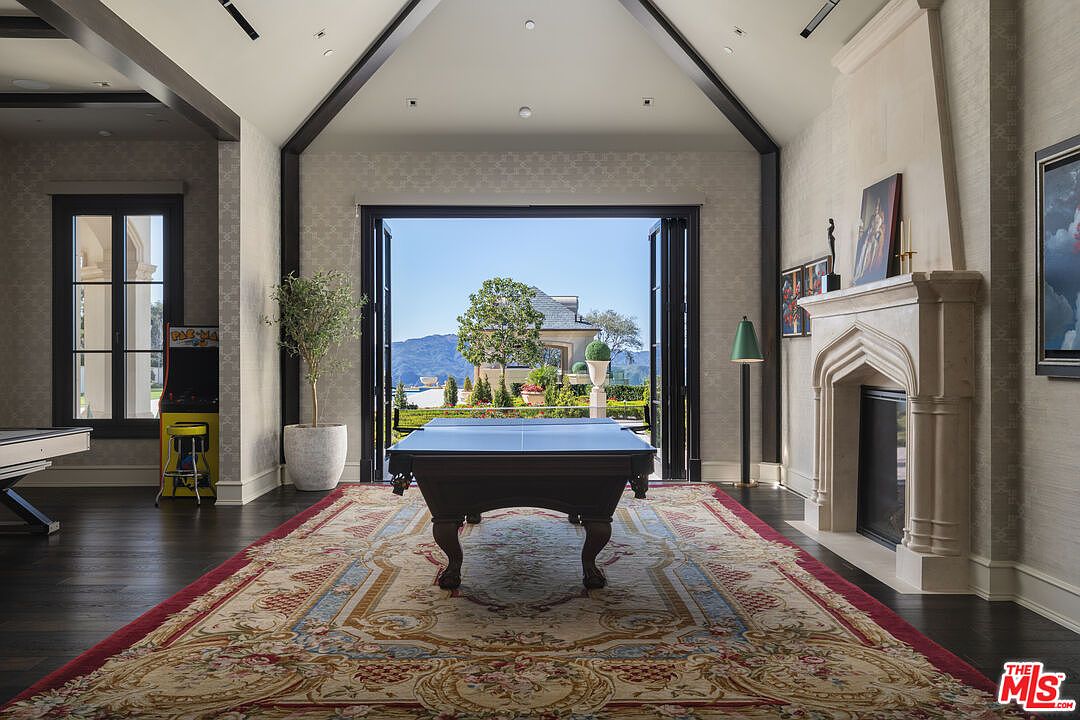 This interior shot showcases a luxurious living room featuring a pool table as a focal point, complemented by a patterned rug and a stone fireplace. Large open doors provide a view of the exterior landscape, enhancing the room's spacious feel and connection to the outdoors. The room is decorated with a mix of modern and traditional elements, creating an elegant and inviting atmosphere.