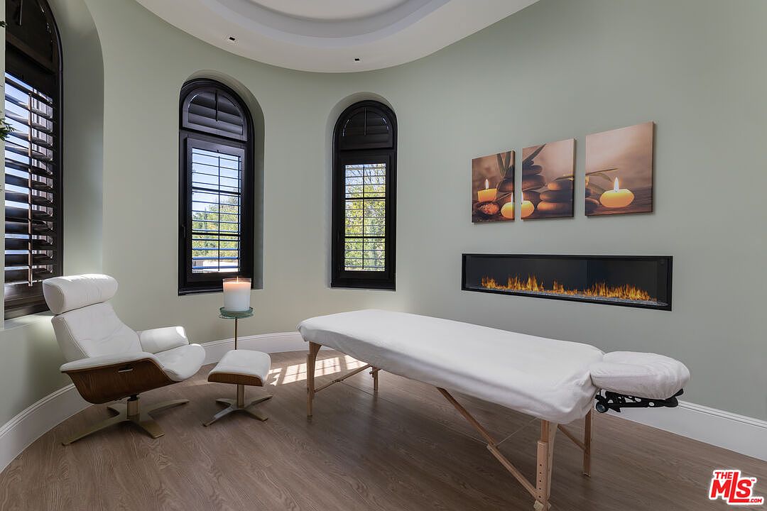 This interior shot showcases a serene spa or massage room, featuring a massage table with white linens, a modern lounge chair with ottoman, and a linear fireplace beneath artwork depicting candles and stones. The room is painted in a calming light green, with arched windows providing natural light, creating a tranquil and inviting atmosphere.