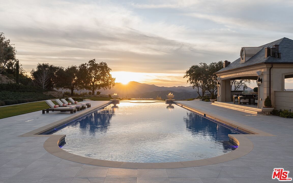This image showcases a luxurious infinity pool at sunset, reflecting the sky and surrounding mountains. The pool is surrounded by a spacious patio with lounge chairs and a covered outdoor living area, creating an inviting and serene atmosphere. The architecture of the adjacent building suggests a high-end estate, emphasizing the property's exclusivity and appeal.