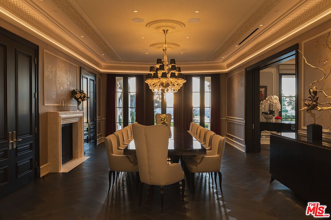 This is a luxurious dining room featuring a long, dark wood table surrounded by elegant beige chairs. A grand chandelier hangs above the table, providing warm lighting. The room is decorated with sophisticated wall paneling, a fireplace, and large windows that offer a view of the outside, creating an opulent and inviting atmosphere.