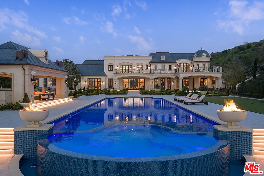 This image showcases a luxurious backyard featuring a large, uniquely shaped swimming pool with integrated spa. The pool is surrounded by a spacious patio area with lounge chairs and fire bowls, leading up to a grand, classically styled mansion with multiple balconies. The scene is set against a backdrop of lush greenery and a clear sky, creating an inviting and opulent atmosphere.