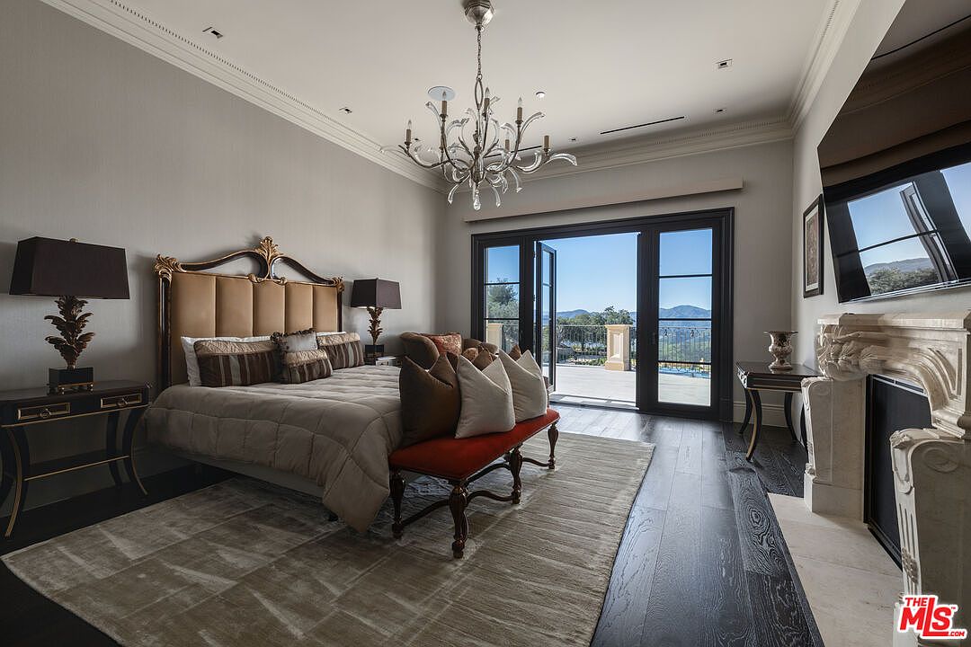 This is a luxurious primary bedroom featuring a large bed with an upholstered headboard, a decorative chandelier, and dark hardwood floors. A set of glass doors leads to an outdoor balcony with scenic views. The room also includes a fireplace and elegant side tables with lamps, creating a sophisticated and comfortable atmosphere.