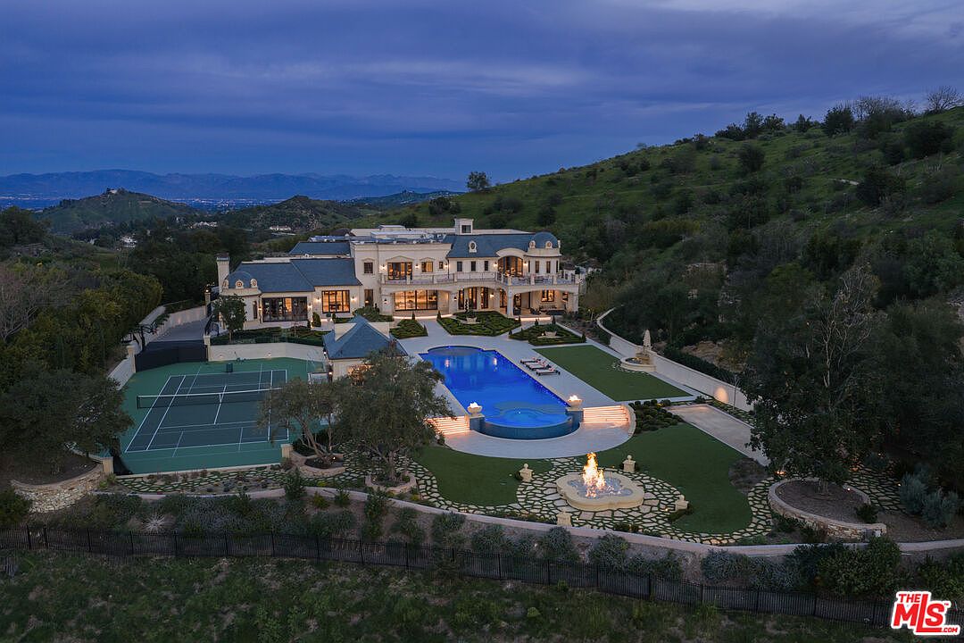 This aerial view showcases a luxurious estate at dusk, featuring a grand mansion with a swimming pool, tennis court, and meticulously landscaped grounds. The property is nestled on a hillside, offering stunning views of the surrounding landscape and mountains in the distance, creating an impression of exclusivity and serenity.