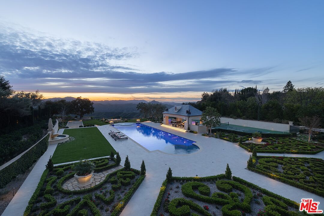 This aerial view showcases a luxurious estate featuring a large swimming pool with a pool house, meticulously manicured gardens with intricate designs, and a tennis court. The property is surrounded by lush greenery and offers stunning views of the distant mountains, creating a serene and private oasis. The overall impression is one of elegance and sophistication.