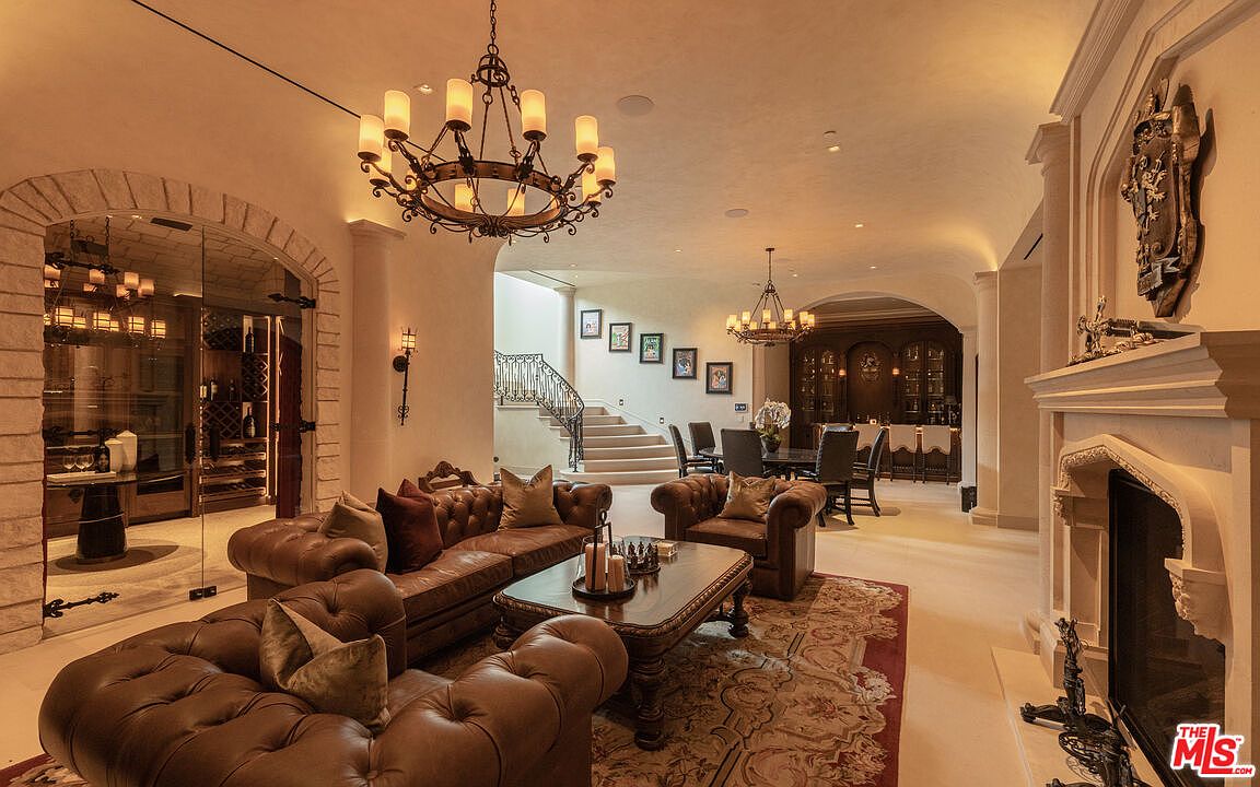 This is an interior shot of a luxurious living room featuring a grand staircase, a wine cellar behind a glass wall, and a formal dining area in the background. The room is furnished with plush leather sofas and a decorative rug, illuminated by ornate chandeliers, creating an opulent and inviting atmosphere.