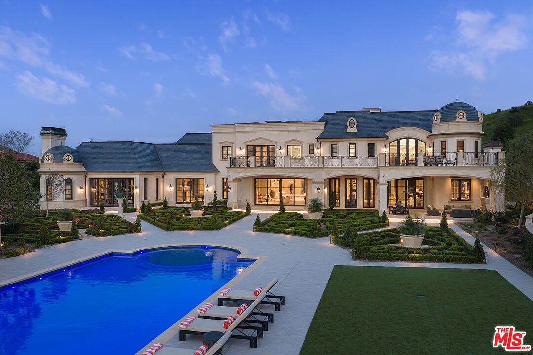 This image showcases the rear exterior of a luxurious estate, highlighting a stunning pool and meticulously landscaped grounds. The architectural style is elegant and refined, featuring a grand building with multiple balconies and large windows. The pool area includes lounge chairs, and the surrounding garden is manicured with geometric hedges and lush greenery, creating a serene and upscale ambiance.