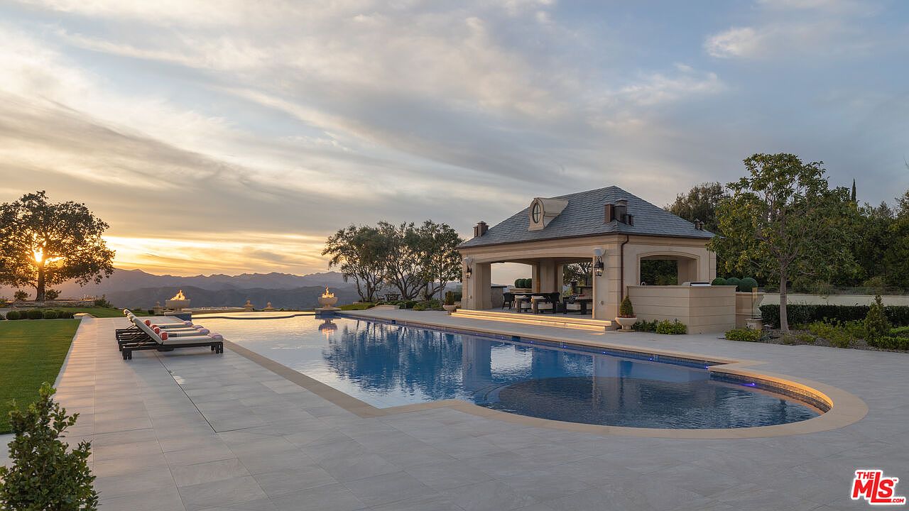 This is a stunning exterior shot of a luxurious pool and spa area at sunset. The long, rectangular pool features a curved end and is surrounded by a spacious patio. A covered outdoor living area with a dining table sits at the end of the pool, and fire bowls add a touch of elegance. The scene is framed by lush landscaping and a picturesque mountain backdrop, creating a serene and inviting atmosphere.