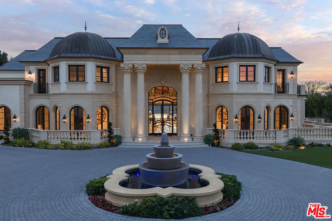 This is a front exterior view of a grand, luxurious mansion. The architecture features symmetrical design with domed towers, classical columns, arched windows, and a large ornate entrance. A circular driveway with a tiered fountain in the center adds to the property's opulence and curb appeal.