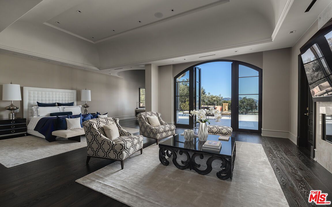 This is a luxurious primary bedroom featuring a large bed with a tufted headboard, dark wood floors, and a seating area with patterned armchairs and a black ornate coffee table. The room is bathed in natural light from the large arched windows that lead to a balcony, offering a serene and elegant atmosphere. A fireplace is visible on the right side of the image.