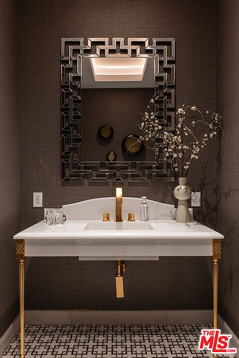 This is a stylish guest bathroom featuring a white marble countertop with gold legs and fixtures. A decorative mirror hangs above the sink, complemented by modern wall decor and a vase with floral accents. The patterned floor adds a touch of sophistication to the space.