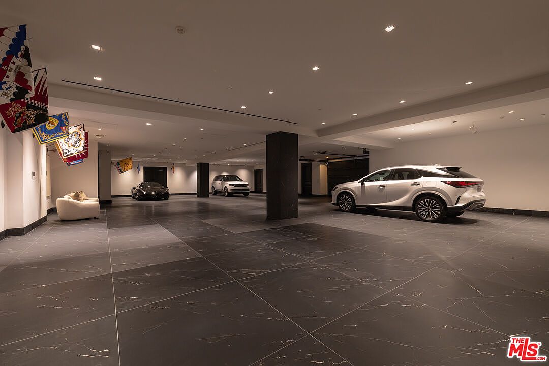 This is a spacious, modern garage featuring a dark tile floor and recessed lighting. Three cars are parked within the garage, and decorative flags hang on the left wall. The garage appears to be well-lit and designed with a minimalist aesthetic.