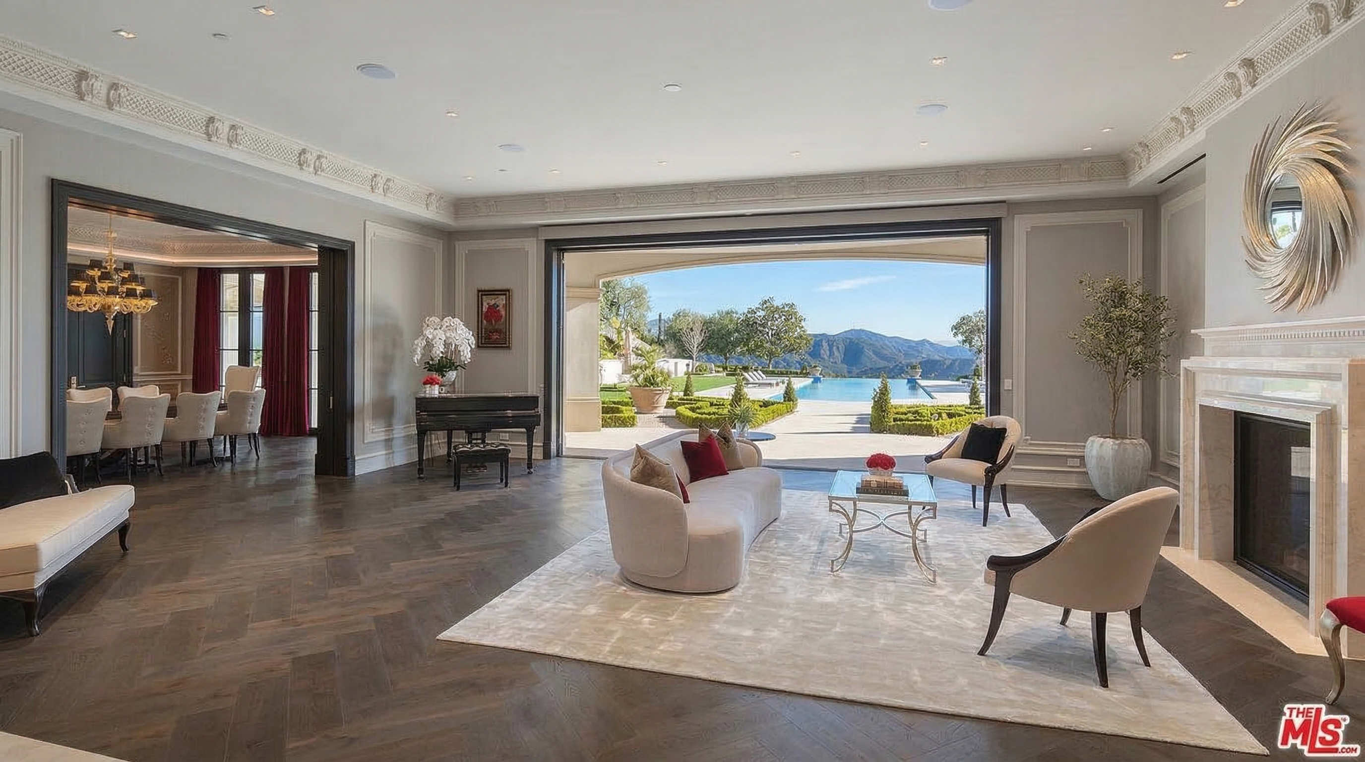 This elegant and spacious living room features a sophisticated open-concept design that transitions seamlessly to a scenic outdoor pool area. The room is anchored by a plush, curved sofa and high-end furniture set atop a large area rug, complemented by warm hardwood flooring and decorative crown molding. A grand piano and a fireplace framed by a distinctive circular wall mirror add a sense of luxury and depth to the airy, bright space.
