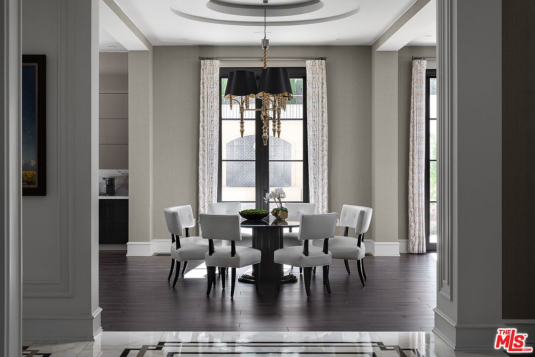 This is an interior shot of a dining room featuring a round table with six chairs, illuminated by a chandelier with black shades. The room has a neutral color palette with patterned curtains framing the windows, and dark hardwood floors add a touch of elegance. The space is well-lit and appears sophisticated, suitable for formal dining.