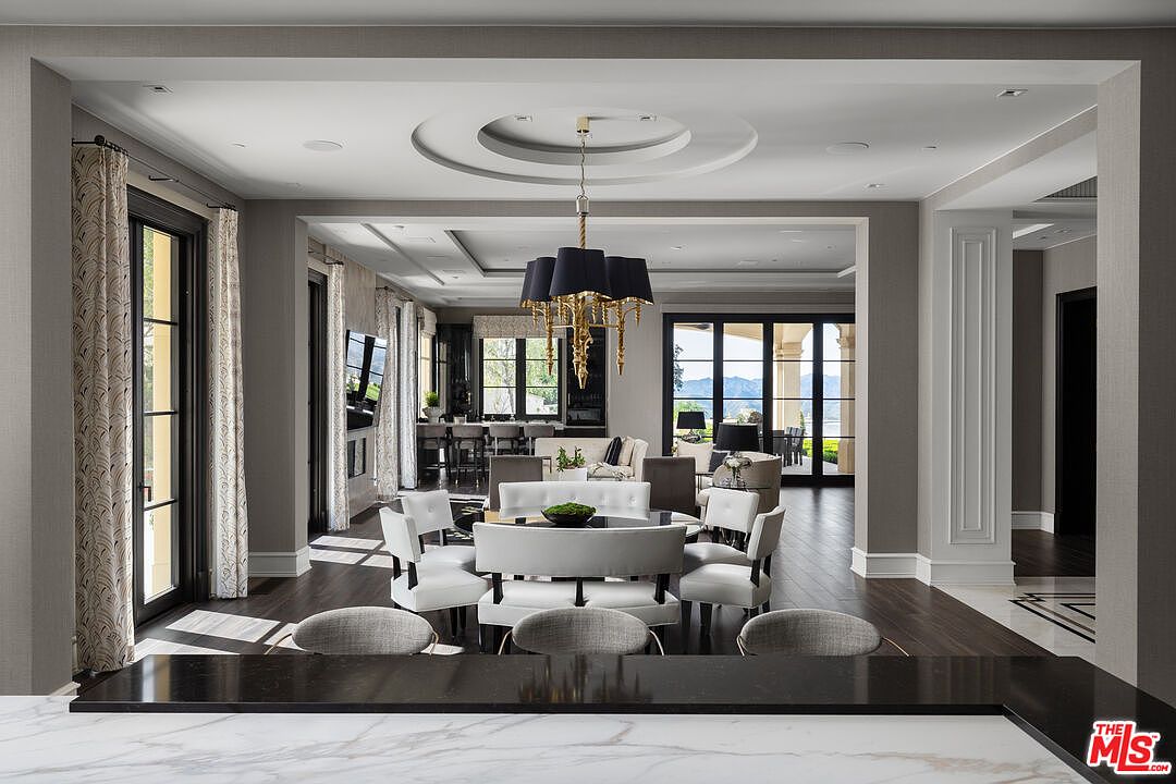 This interior shot showcases a luxurious dining area with a round table surrounded by white chairs and a unique chandelier. The space is open and flows into other living areas, with large windows providing natural light and views. Dark wood flooring and neutral wall colors contribute to the elegant and sophisticated ambiance.