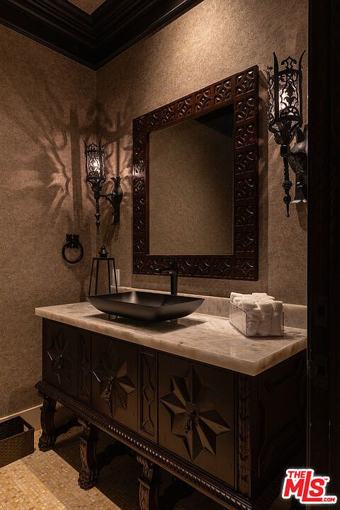 This is a powder room featuring a dark wood vanity with a marble countertop and a black vessel sink. A decorative mirror hangs above the sink, flanked by ornate sconces. The walls are textured, and the overall style is elegant and somewhat gothic.