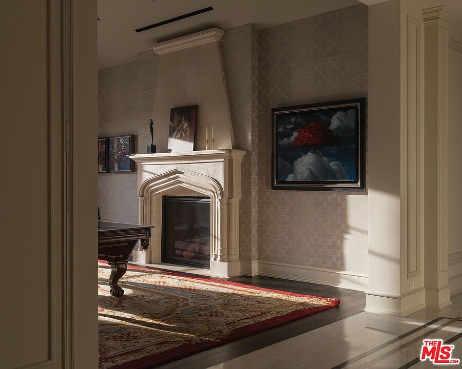 This interior shot showcases a sophisticated living room featuring a stone fireplace with an ornate mantel, complemented by artwork on the walls. A patterned rug adds warmth to the dark wood flooring, while natural light streams in, highlighting the architectural details and creating an inviting atmosphere. A glimpse of a pool table adds a touch of leisure.