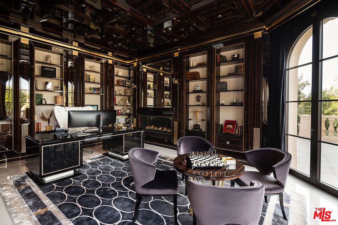 This luxurious office features dark wood built-in bookshelves with gold accents, a glossy black desk, and plush purple chairs around a chess table. The room is illuminated by natural light from a large arched window, and a modern fireplace adds warmth. The overall impression is one of sophistication and opulence, perfect for a high-end real estate listing.