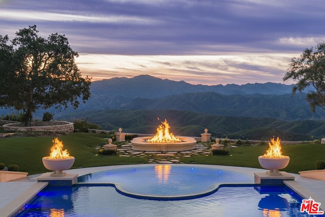 This image showcases a luxurious outdoor space featuring a uniquely shaped pool with vibrant blue water, complemented by two fire bowls on either side. A central fire pit with a stone base adds warmth and ambiance, set against a backdrop of rolling hills and a dramatic sunset sky. The scene exudes elegance and tranquility, perfect for upscale outdoor living.