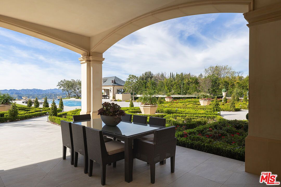 This image showcases a luxurious outdoor patio area, featuring a dining table set for six under a covered archway. The patio overlooks a meticulously manicured garden with geometric hedges and potted plants, extending towards a swimming pool and distant mountains. The scene exudes elegance and tranquility, highlighting the property's outdoor living space.