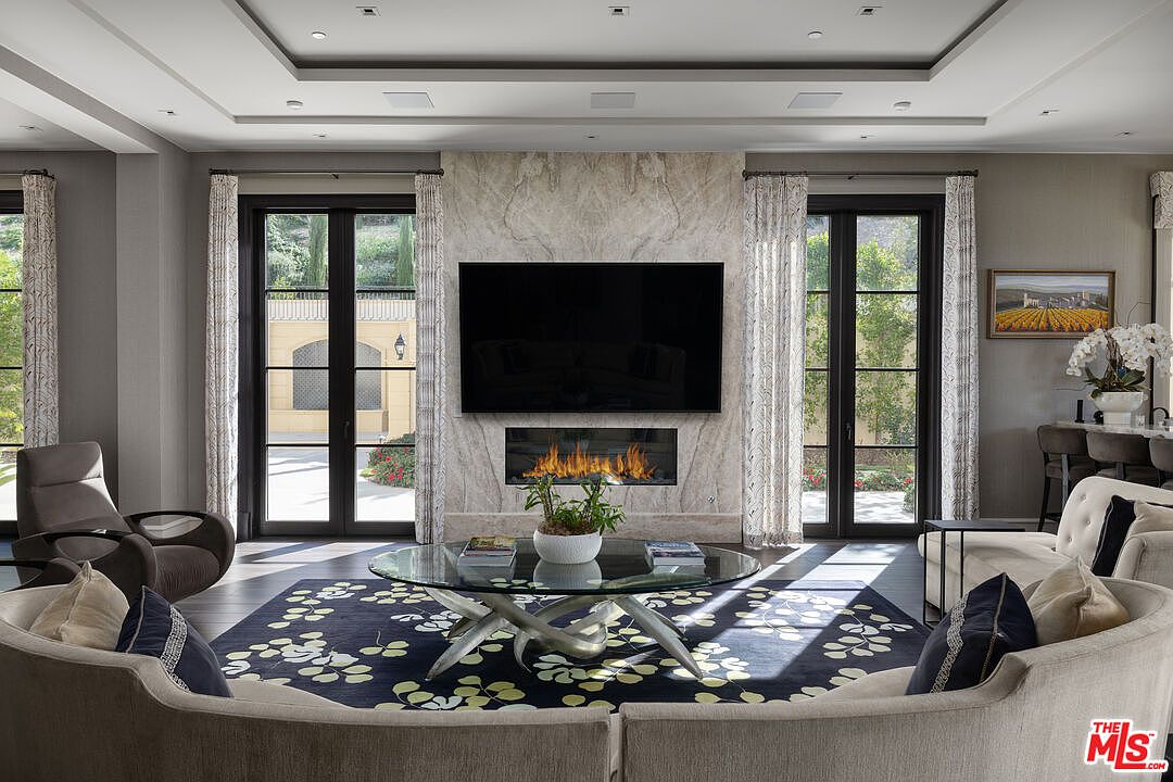 This is a beautifully designed living room featuring a modern fireplace with a large television mounted above it. The room is decorated with neutral tones, complemented by a dark blue floral rug and elegant curtains framing the doors to the outside. The furniture is arranged to create a comfortable and inviting space, perfect for relaxation and entertainment.