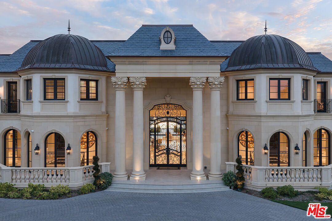 This is a grand front view of a luxurious mansion, showcasing its impressive architectural design. The facade features elegant columns, arched windows, and a stately entrance with ornate iron gates. The manicured landscaping and paved driveway add to the property's curb appeal, creating a strong sense of opulence and sophistication.