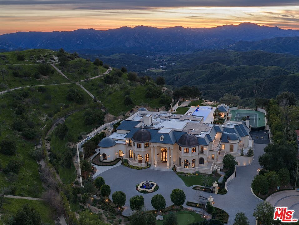 This aerial view showcases a grand estate nestled in a mountainous landscape. The property features a large mansion with a circular driveway, manicured gardens, a tennis court, and a swimming pool. The architectural style is luxurious and impressive, suggesting a high-end real estate offering.