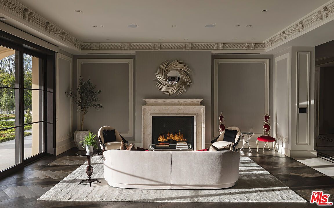 This is an interior shot of a luxurious living room featuring a modern fireplace with a decorative mirror above it. The room is decorated in neutral tones with elegant molding and paneling, and the furniture includes a curved sofa, armchairs, and decorative side tables. Large windows provide natural light and a view of the exterior landscape, creating a sophisticated and inviting atmosphere.