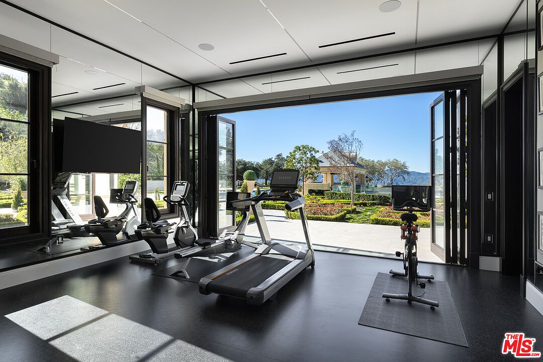 This is a modern home gym featuring state-of-the-art exercise equipment, including a treadmill, stationary bikes, and mirrored walls. Large bi-fold doors open to a beautifully landscaped garden, providing an open-air workout experience. The black flooring and sleek design create a sophisticated and motivating atmosphere.