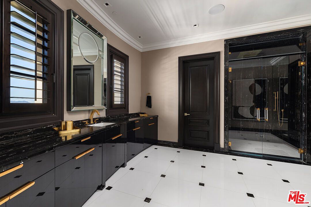 This is a luxurious primary bathroom featuring a long black vanity with gold hardware and a black marble countertop. The floor is white tile with black accents, and a large mirror hangs above the sink. A glass-enclosed shower with black marble accents is visible to the right, and the overall style is modern and elegant.