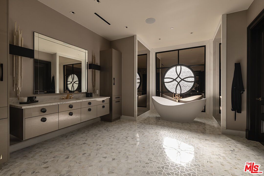 This is a luxurious primary bathroom featuring a freestanding bathtub as a focal point, set against a backdrop of a decorative circular window. The bathroom includes a vanity with drawers and a marble countertop, complemented by a mosaic tile floor. The color palette is neutral, creating a serene and upscale atmosphere.