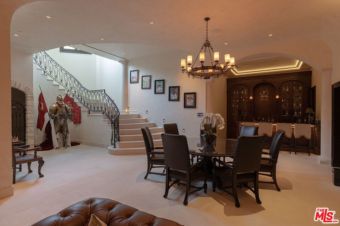 This interior shot showcases a luxurious dining area with a grand staircase and a built-in bar. The dining table is surrounded by elegant chairs, and a chandelier hangs above, providing warm lighting. A suit of armor stands guard near the staircase, adding a unique, historical touch to the space.