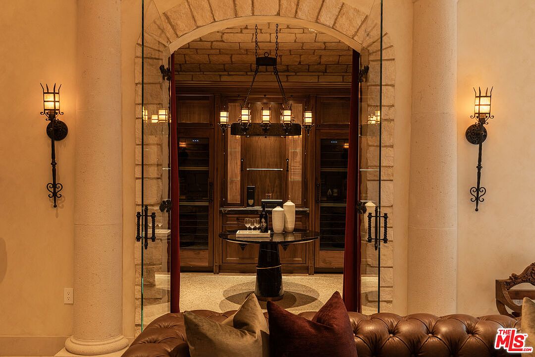 This image showcases a sophisticated wine cellar, featuring a stone archway entrance framed by elegant sconces. Inside, a round table displays wine glasses and decorative vases, set against a backdrop of custom wooden wine storage units. The room exudes a luxurious and inviting atmosphere, perfect for wine enthusiasts.