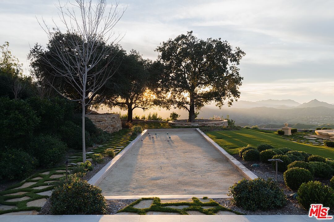 This image showcases a beautifully landscaped yard featuring a bocce ball court as a central element. The court is surrounded by lush greenery, including manicured bushes and trees, with a stone pathway leading to it. In the background, a scenic view of rolling hills and a sunset sky adds to the property's appeal, creating a serene and luxurious outdoor space.