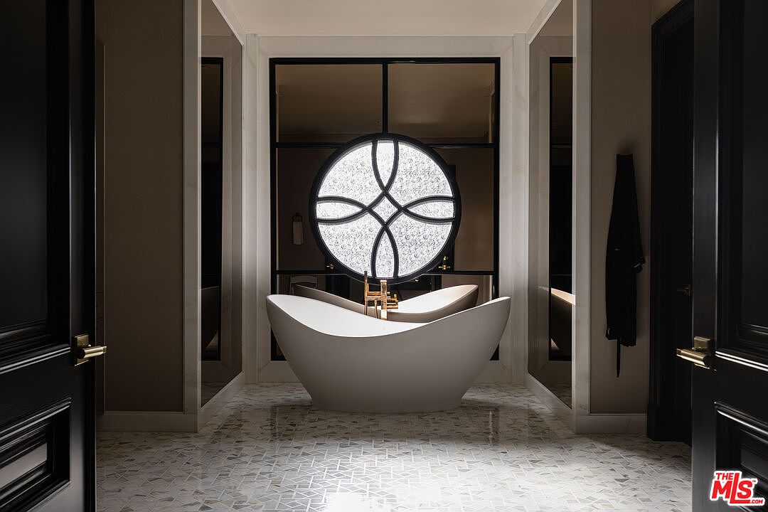This is a luxurious primary bathroom featuring a freestanding white bathtub as the focal point, positioned in front of a decorative circular window. The room is adorned with elegant tile flooring and dark wood doors, creating a sophisticated and spa-like atmosphere. The design emphasizes clean lines and a neutral color palette, enhancing the sense of tranquility and relaxation.