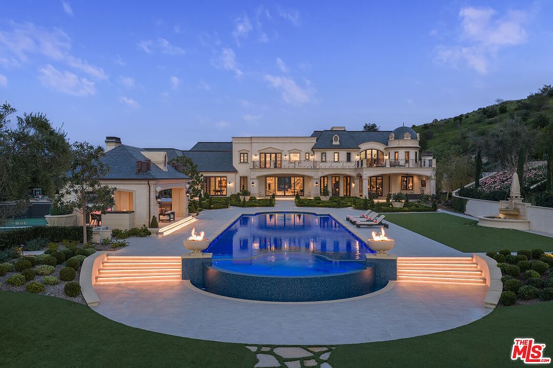 This image showcases the rear exterior of a luxurious estate, highlighting a stunning pool and spa area. The pool features a unique design with integrated fire features, and is surrounded by meticulously landscaped gardens and a spacious patio area with lounge chairs. The grand architecture of the house provides a sophisticated backdrop, emphasizing the property's high-end appeal and outdoor entertainment possibilities.