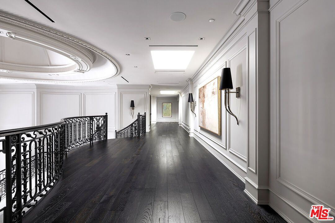 This is an interior shot of a luxurious hallway featuring dark hardwood floors and white paneled walls. The hallway is illuminated by recessed lighting and sconces, and artwork adorns the walls. An ornate black iron railing lines the staircase, adding to the elegant and sophisticated ambiance of the space.
