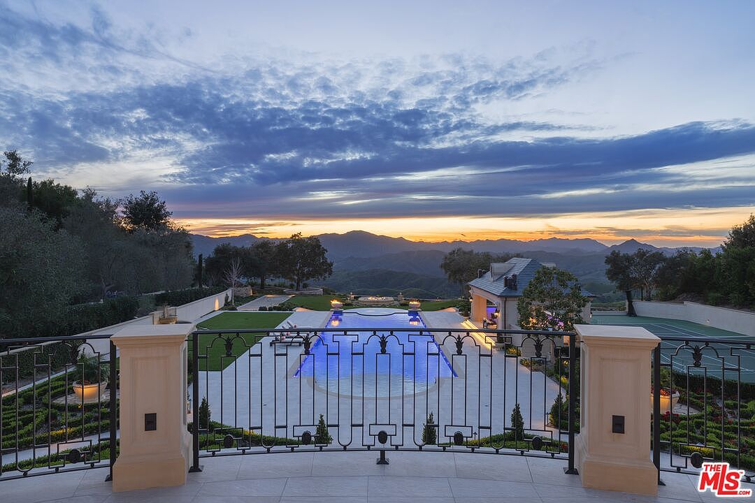 This exterior shot showcases the rear of a luxurious property at dusk, featuring a stunning infinity pool with illuminated blue water, a tennis court, and meticulously landscaped gardens. The view extends to distant mountains under a dramatic sky, creating a sense of grandeur and tranquility. The image is taken from a high vantage point, likely a balcony, emphasizing the property's elevated position and expansive views.