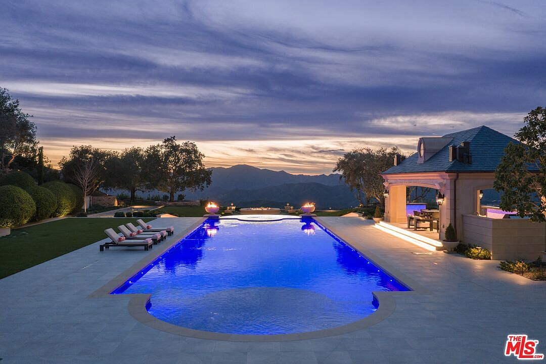 This image showcases a luxurious outdoor pool and spa area at dusk. The rectangular pool features vibrant blue lighting, and is flanked by lounge chairs on one side and a covered outdoor living space on the other. In the background, mountains are visible under a dramatic sky, creating a serene and upscale ambiance.