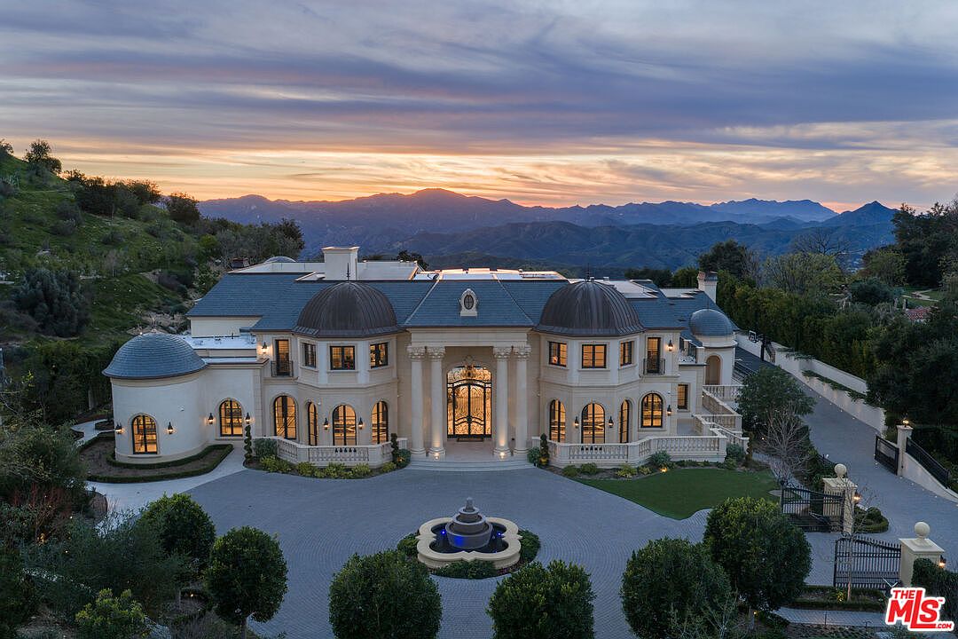 This is a stunning front view of a grand estate, showcasing its impressive architecture with symmetrical design, domed towers, and a stately entrance framed by columns. The property features a circular driveway with a central fountain, manicured landscaping, and a backdrop of rolling hills and a vibrant sunset, creating an overall impression of luxury and elegance.