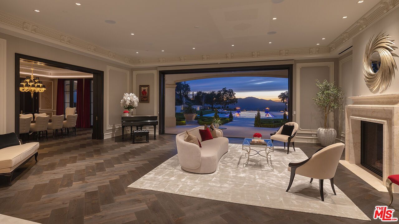 This is an interior shot of a luxurious living room featuring herringbone wood floors, a neutral-toned curved sofa, and two accent chairs arranged around a glass coffee table on a light-colored rug. A grand piano sits near a doorway leading to a dining room, while a large sliding glass door opens to an outdoor pool area with a view of mountains at sunset. A fireplace with a decorative mirror above it adds a touch of elegance.