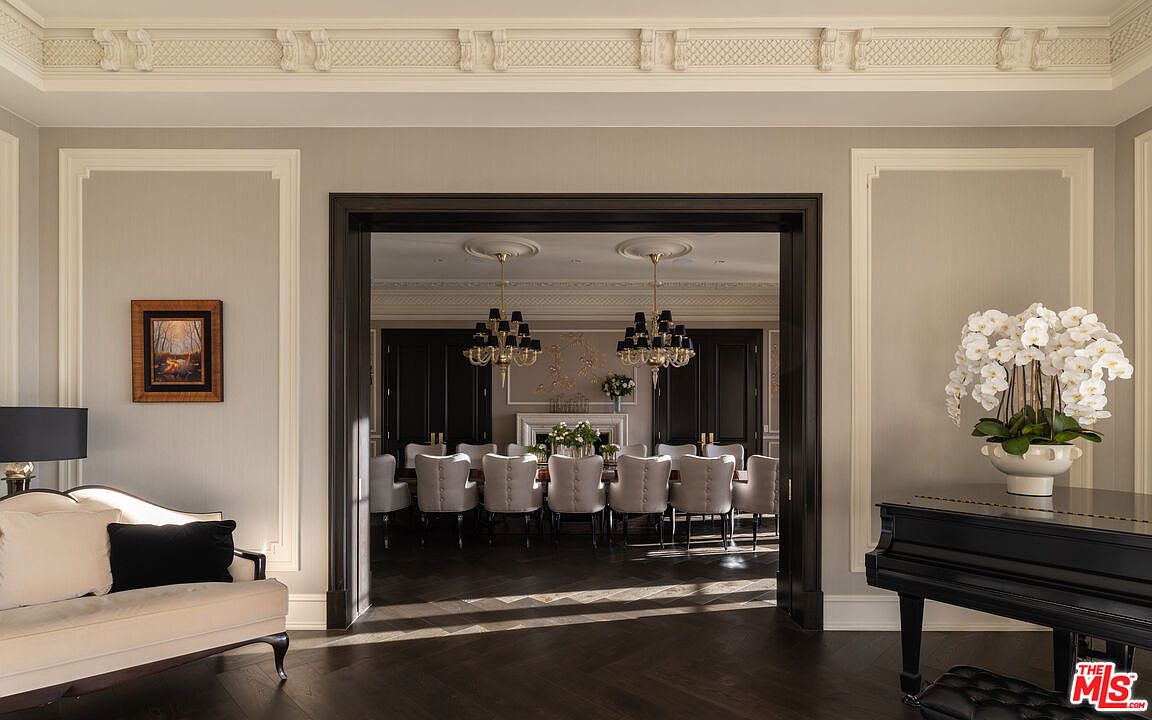 This image showcases an elegant dining room viewed through a doorway from an adjacent living space. The dining room features a long table with multiple chairs, illuminated by two chandeliers, and a fireplace with decorative accents. The living room has a sofa and a piano, suggesting a luxurious and sophisticated home.